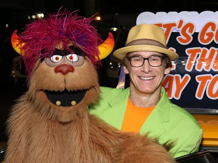 Man with yellow top hat and black-framed glasses wearing a bright green suit holds a monster puppet