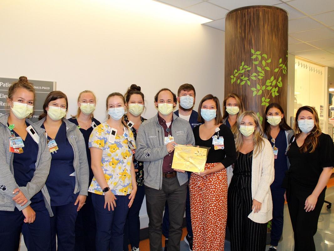 A dozen members of the Pediatric Intermediate Care Unit at Penn State Health Children’s Hospital, wearing masks and scrubs, stand together in a hospital corridor while displaying an award certificate.