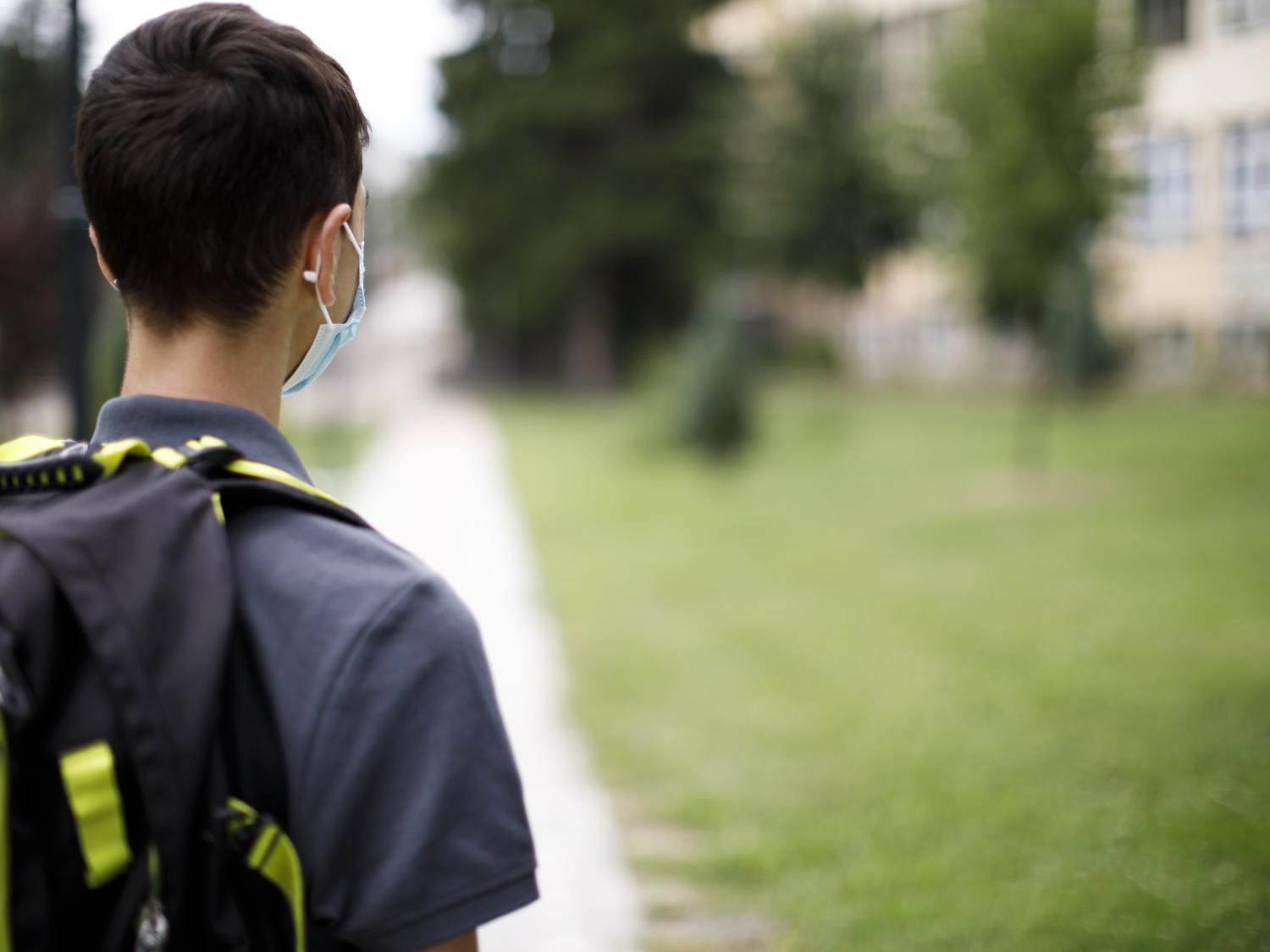 A boy wearing a mask and backpack stares at a school building off in the distance.
