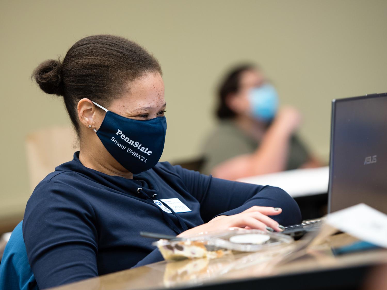 Woman student wearing a mask who is working at a desk on her laptop in a classroom. 