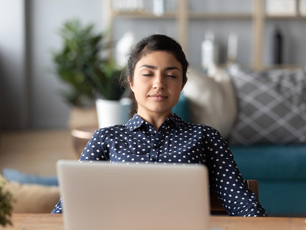A woman sits in front of her laptop with her eyes closed while meditating.