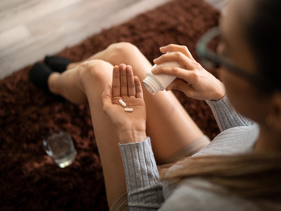 A young woman pours pills from a bottle into her hand. A glass of water sites on the floor in front of her. 