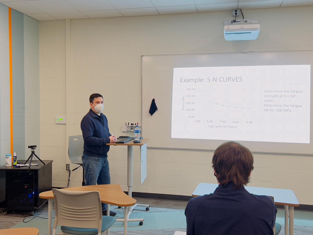 Man standing at front of room behind a podium. A projector projects a chart on a white board. In the forefront of the photo there is a student looking at the man.
