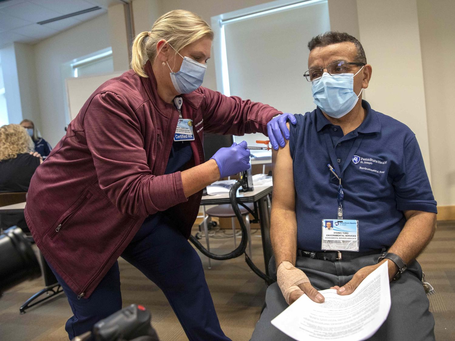 A nurse manager leans over slightly as she administers a vaccine to a man who is seated in a chair.
