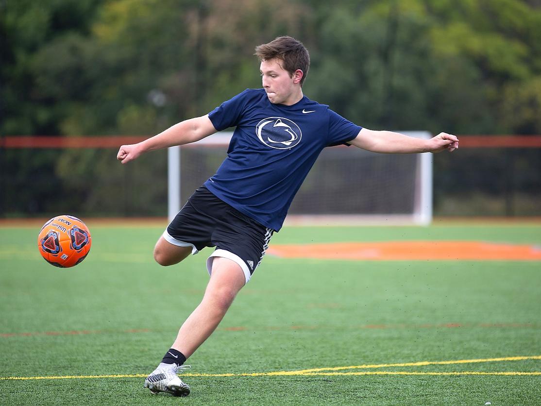Brayden Sunho gets ready to kick a soccer ball that is in mid-air. Brayden, who is wearing shorts and a shirt with a Penn State Nittany lion logo on the front, is running on a soccer field marked with goal lines.