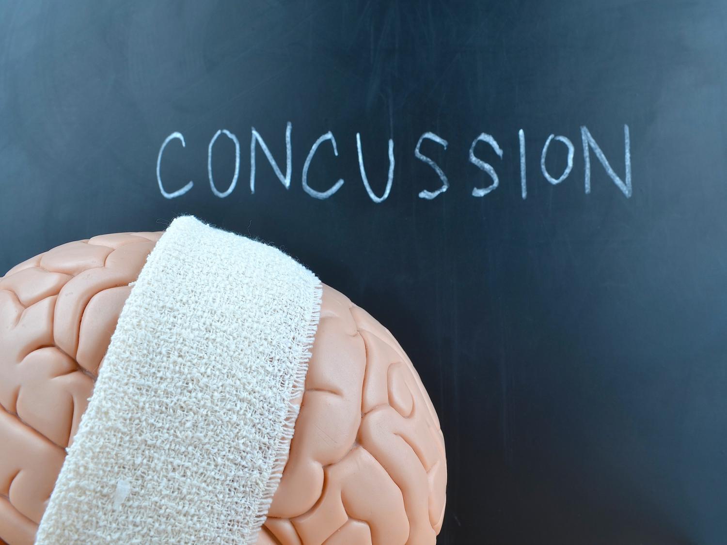 An illustration shows a brain draped with an athletic towel. Behind it on a chalkboard is written the word “concussion.”