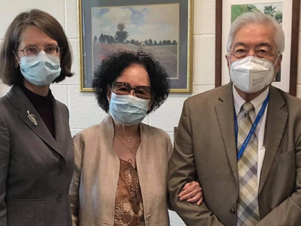 Three people stand next to one another in a room, all wearing face masks. One woman wears a brown suit, and another woman wears a brown jacket. She has her arm linked with a man who is wearing a brown suit and blue checkered tie. 