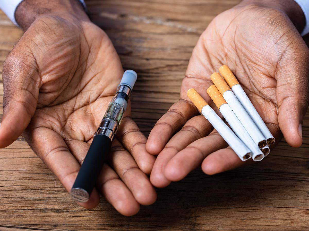 A person holds out their hands. The right hand contains an electronic cigarette, while the left holds five cigarettes.