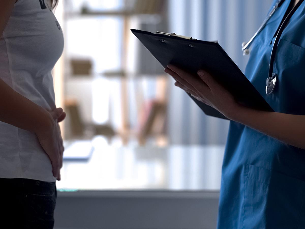 A woman holds her abdomen while a medical professional wearing scrubs standing across from the woman holds a clipboard.