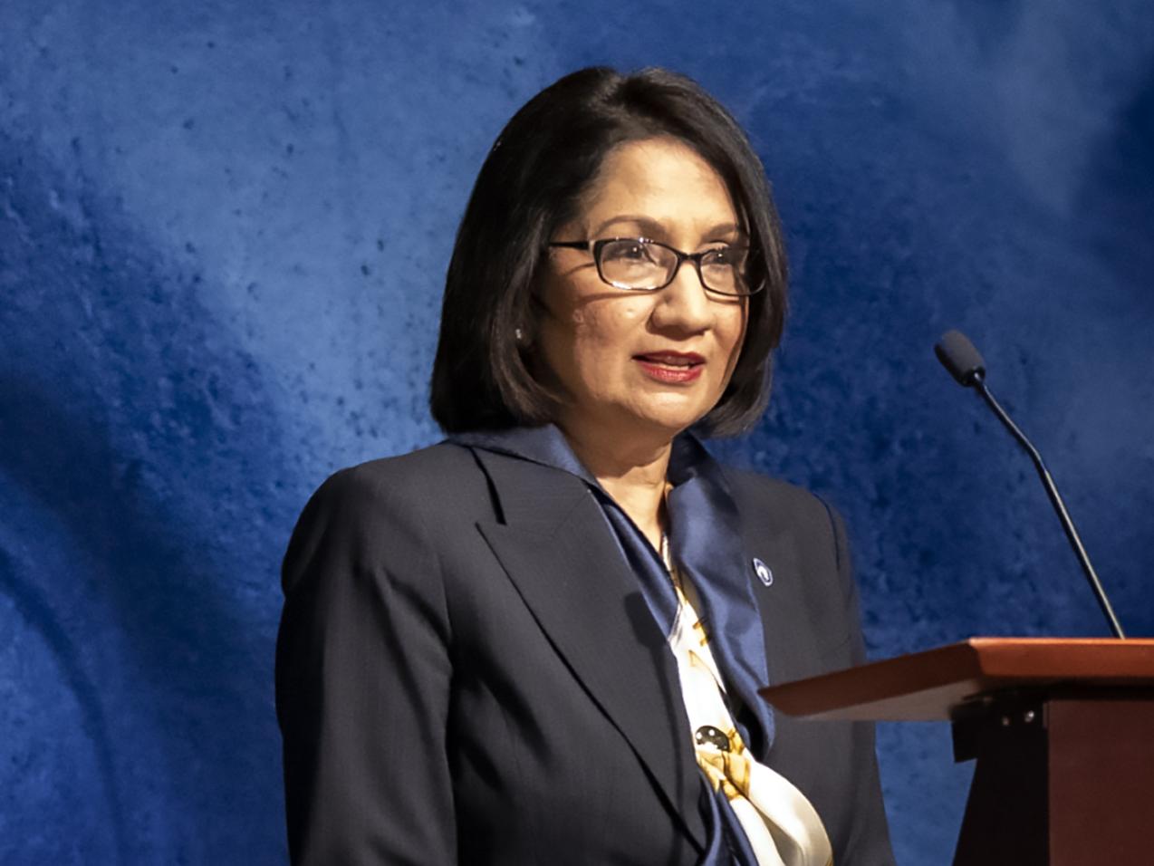 Neeli Bendapudi standing at a podium with a blue backdrop behind her.