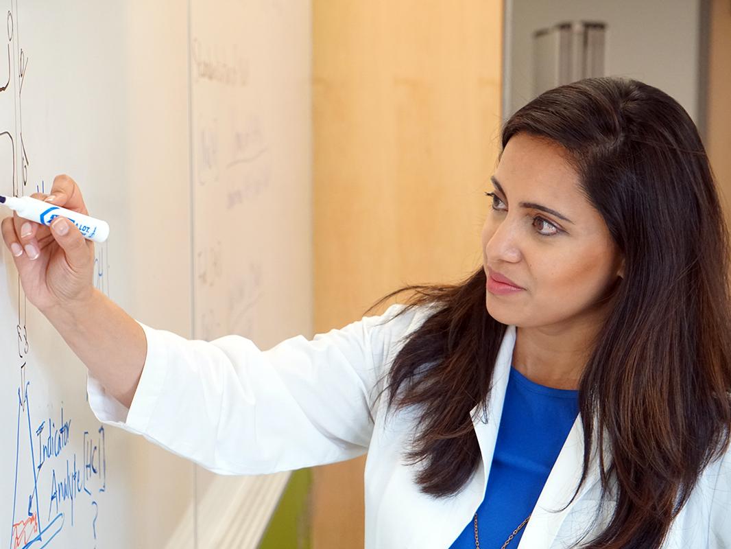 Woman with long brown hair in white lab coat drawing on a white board with a marker.