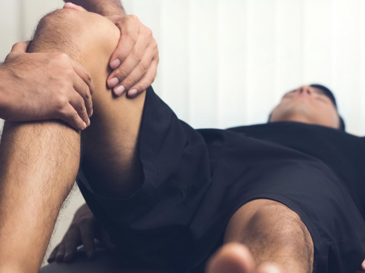 A patient lays flat on a treatment table with his right knee bent. A provider maneuvers the knee with both hands.