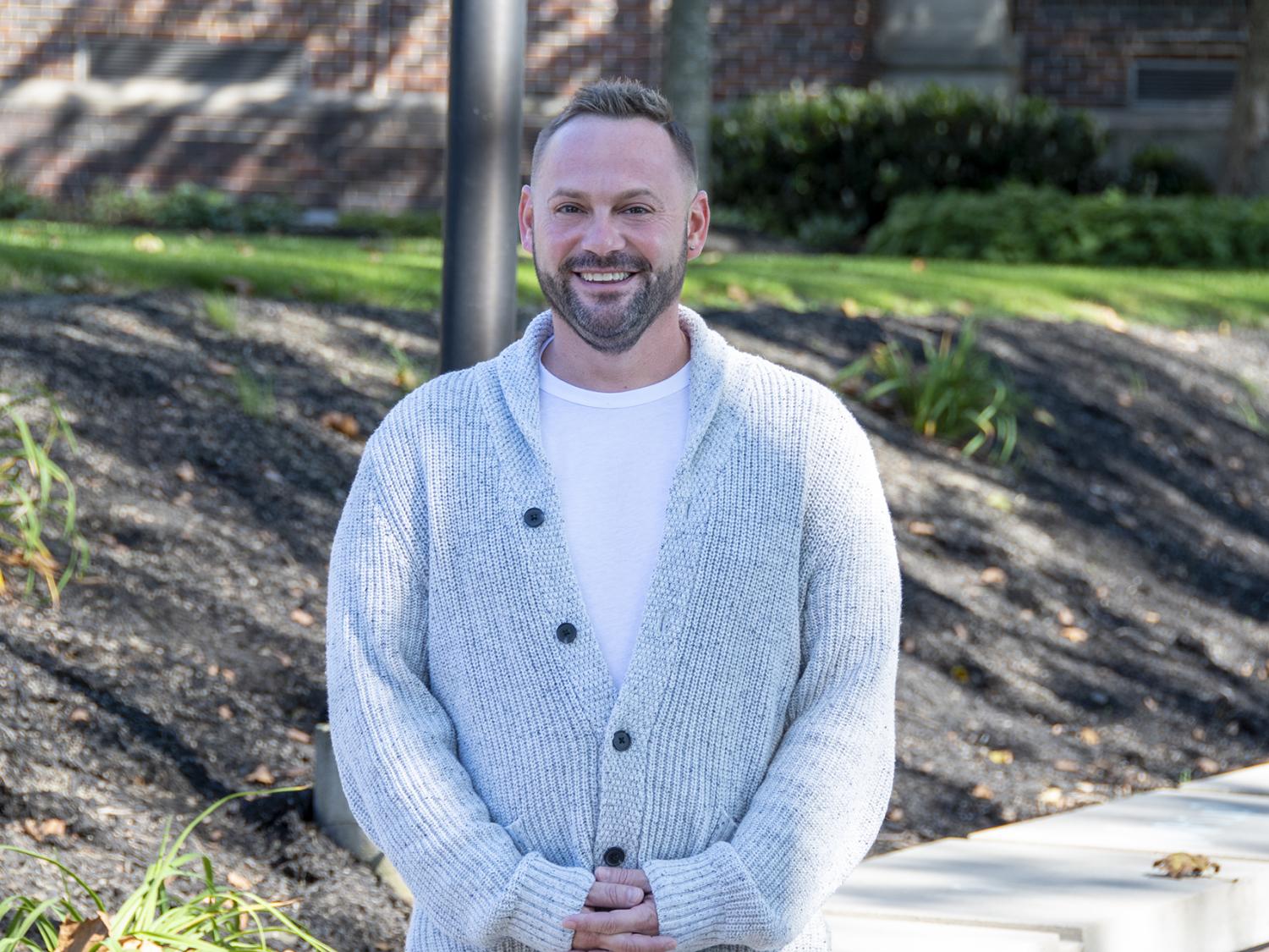 Smiling man wearing a white shirt and cardigan stands with hands clasped in front of him.