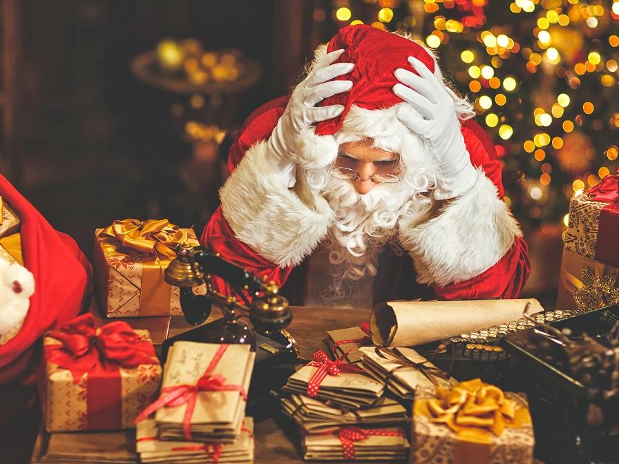‘Santa Claus’ rests his elbows on a desk and his head on his hands. The desk is covered with presents and letters. A Christmas tree is in the background, out of focus.