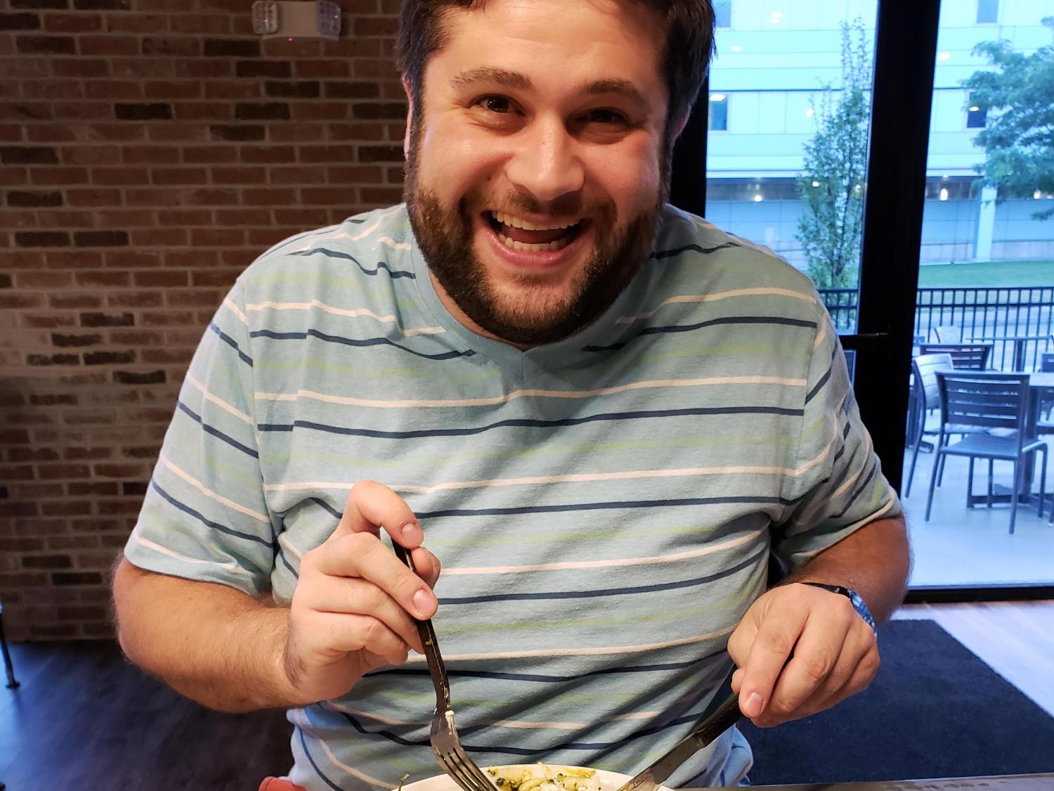 Alex Goodstein smiles for the camera while holding a fork and knife before taking his first bite of a meal