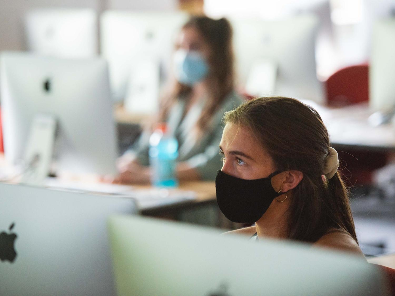 a woman wearing a mask in a classroom sitting at a computer