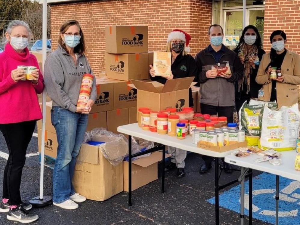 Six people hold food containers near a pile of boxes stationed in a parking space outside a brick building.