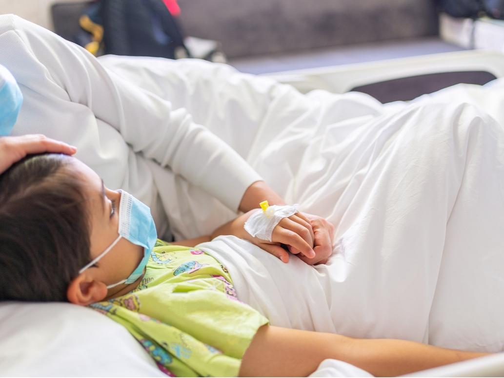 Young mother with protective face mask holding son's hand while lying on the hospital bed with him.