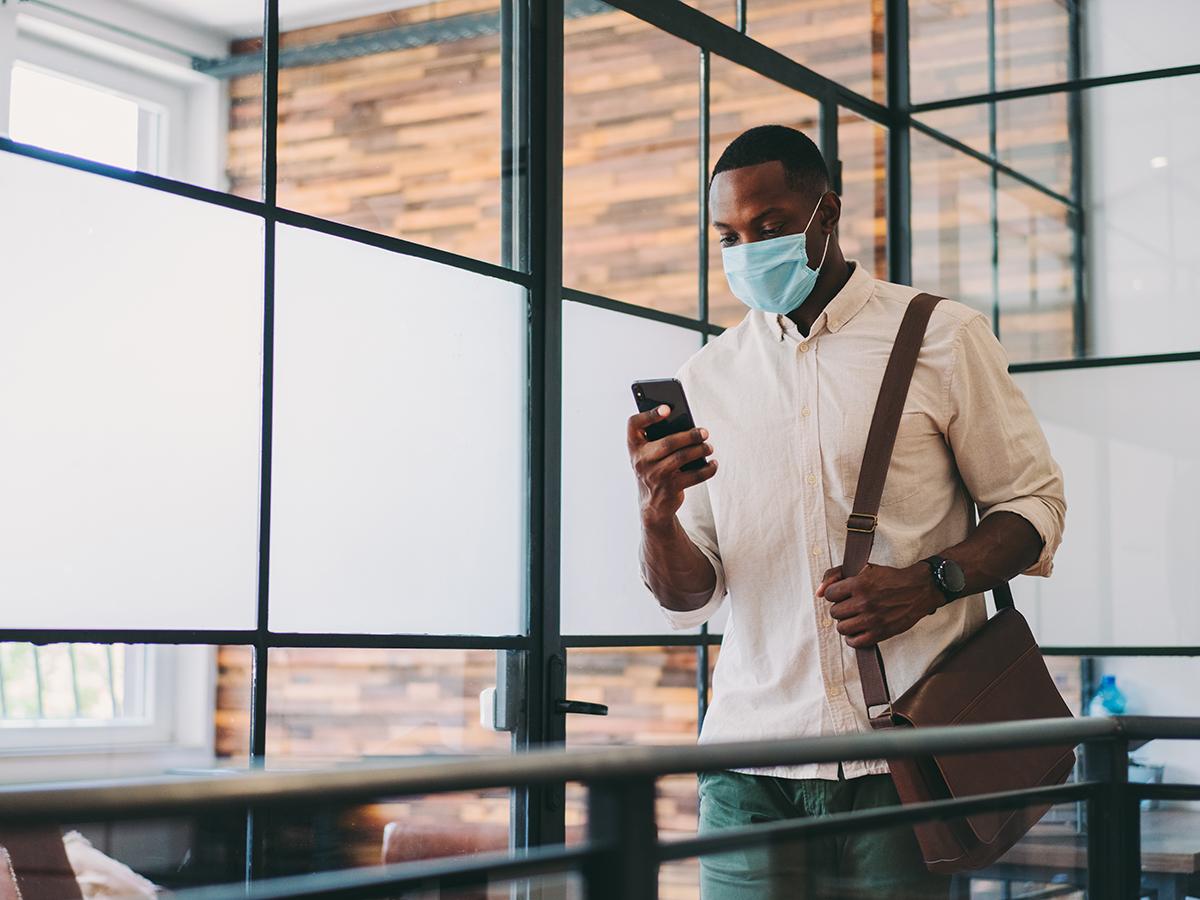 A man wearing a mask checks his phone while walking inside of a building.