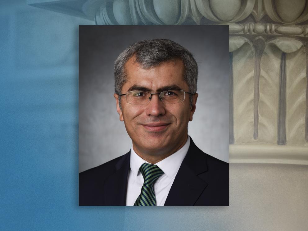 An engineer wearing a green tie, black jacket, and glasses smiles for a portrait on a blue background.