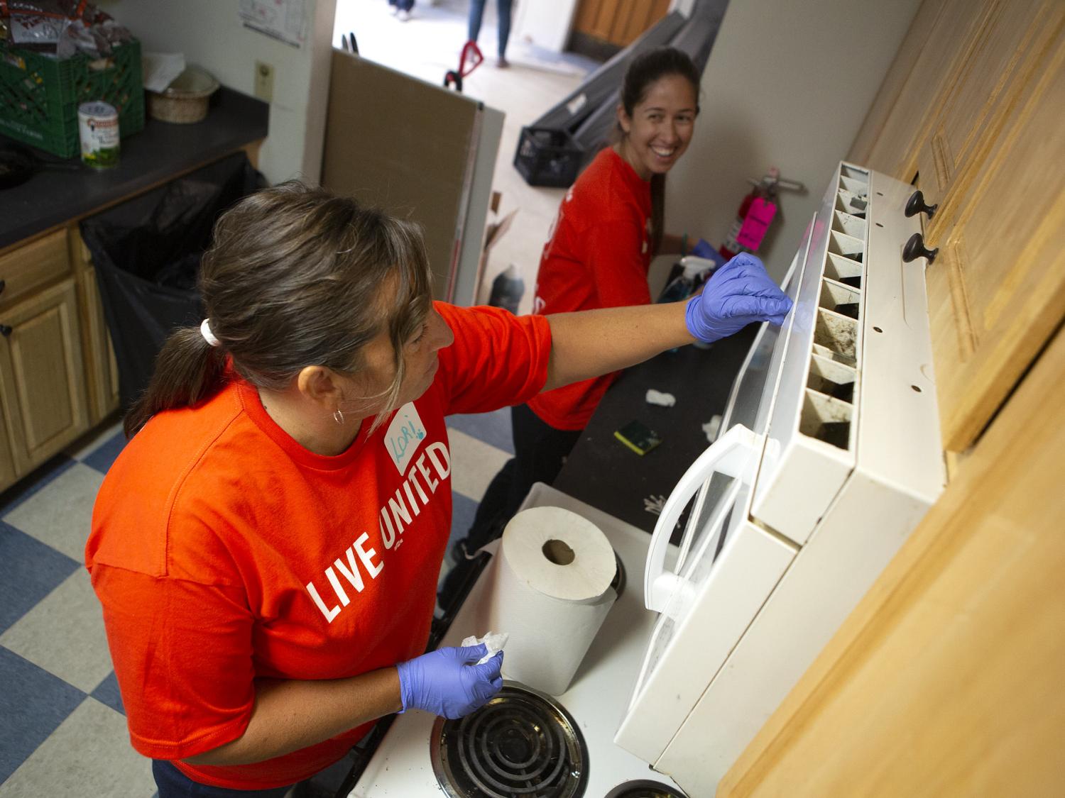 Two women wearing rubber gloves work side-by-side to clean a kitchen in a residential apartment. One looks to the other, smiling.