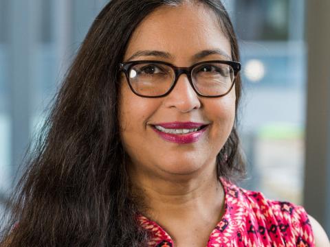 An Indian woman with long dark hair and glasses smiles at the camera. 