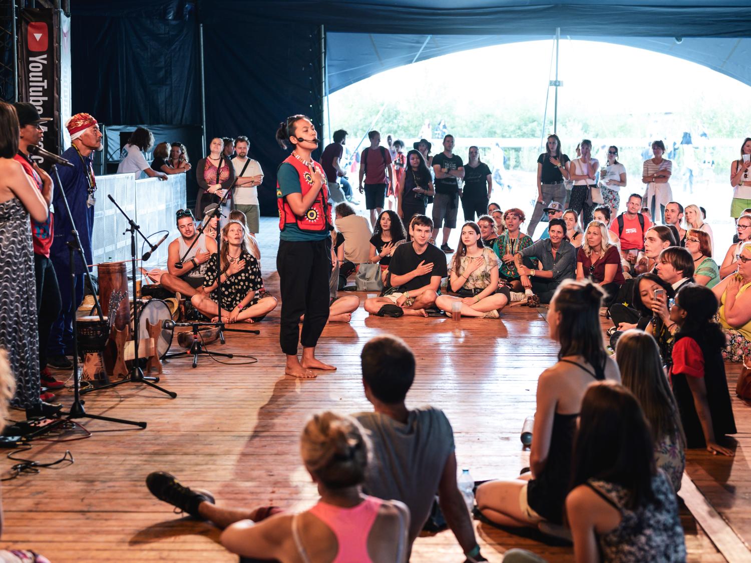 Four people stand in front of microphones and sing in front of a diverse crowd seated in front of them.