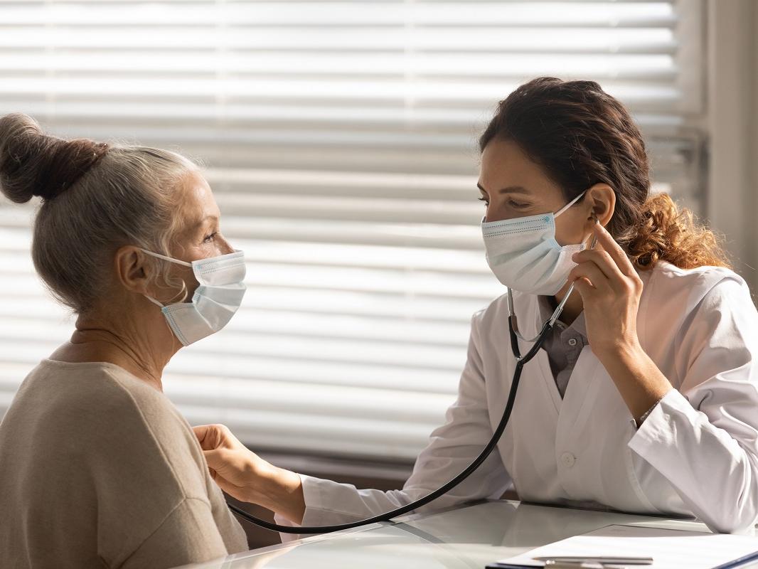 A doctor in a surgical mask hold a stethoscope as she listens to a patient’s heart.