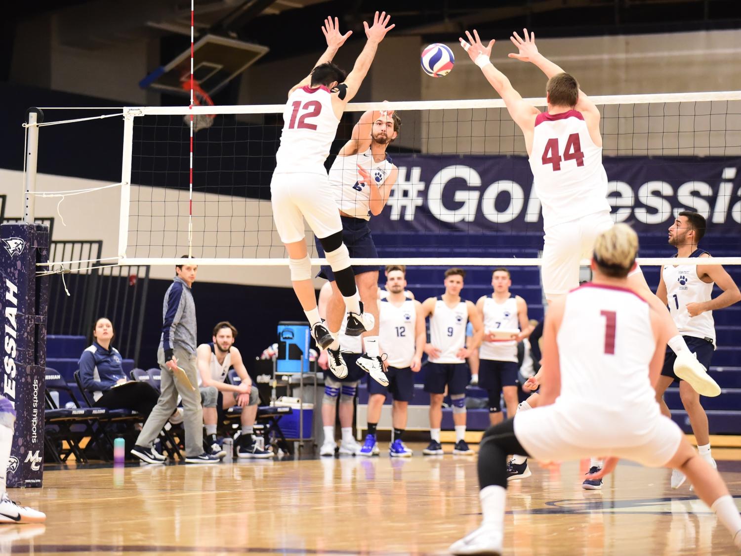 A Penn State Behrend volleyball player jumps to spike the ball over two blocking opponents.