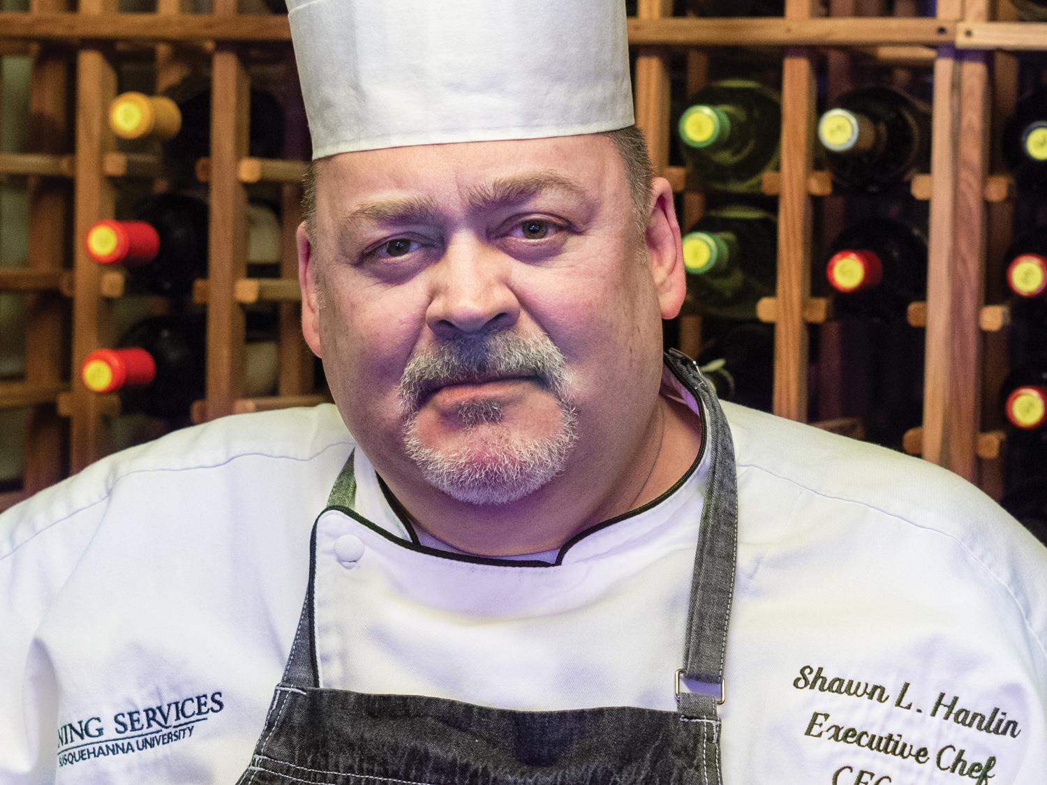 Chef Shawn Hanlin in his white chef coat and hat with racks of wine bottles in the background