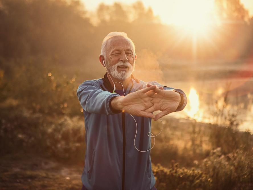 An elderly man stretches his hands before him, he is standing in front of a lake at sunset