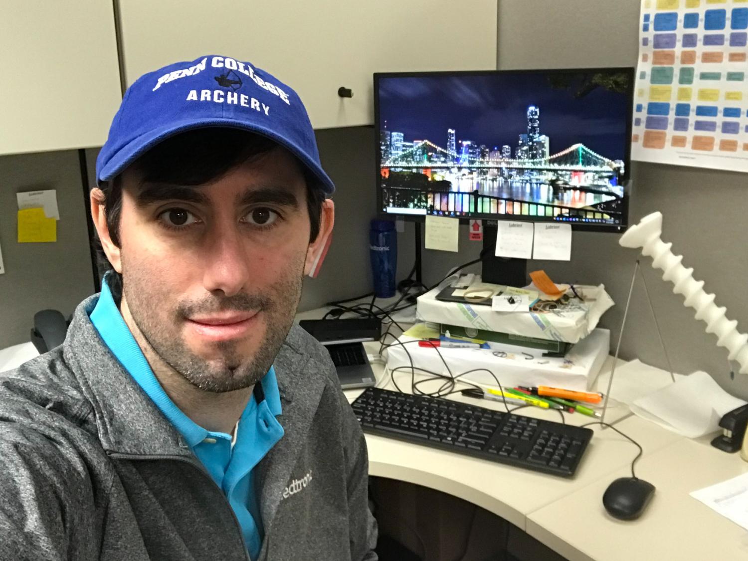 James Fanelli sitting at a desk wearing a blue hat with a computer behind him
