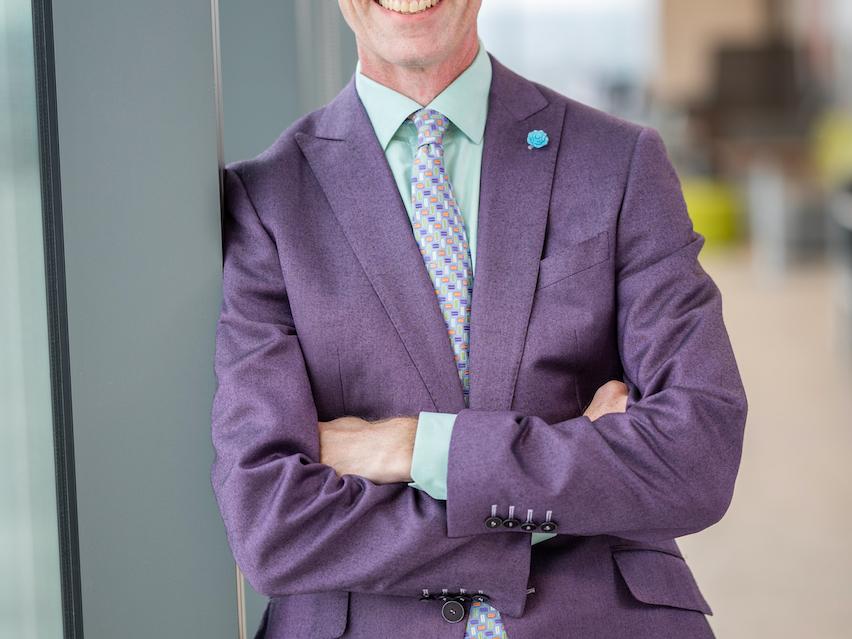 white male in purple suit leaning against window in hallway
