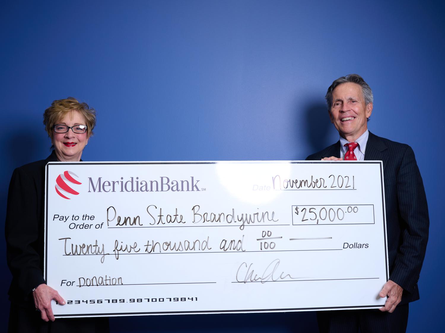 Meridian CEO Chris Annas with Penn State Brandywine Chancellor Marilyn Wells stand in front of a blue wall holding a large white check. 