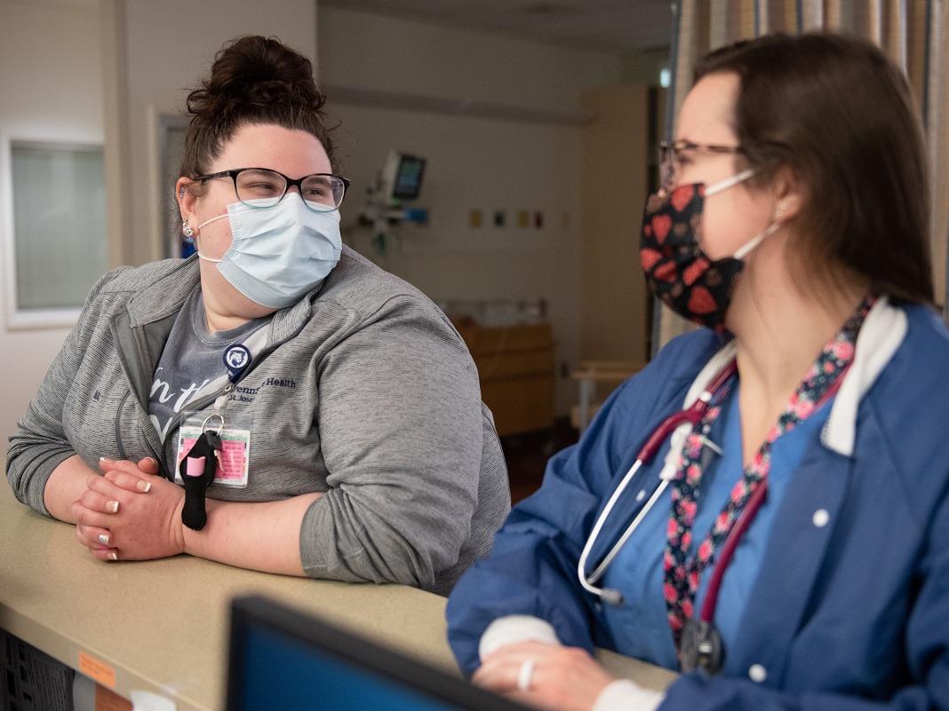 Two women lean on a counter while talking. Both women are wearing glasses and face masks.