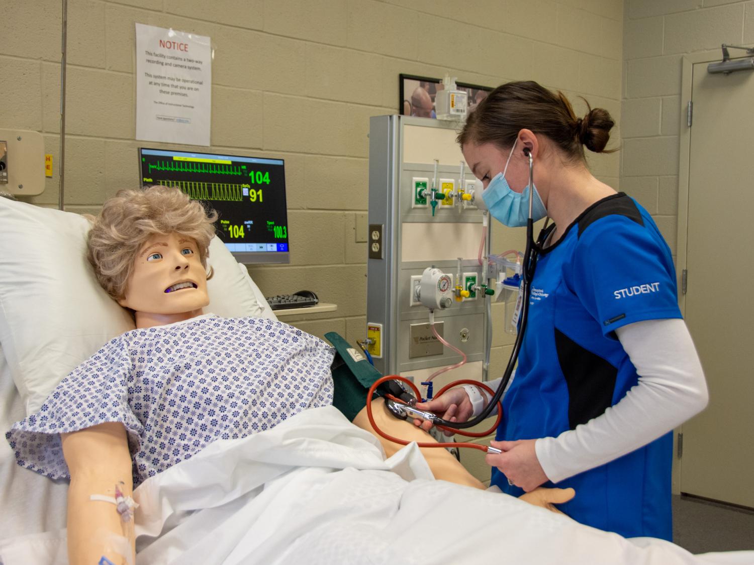 A nursing student wearing blue scrubs interacts with a patient simulator in a hospital room.