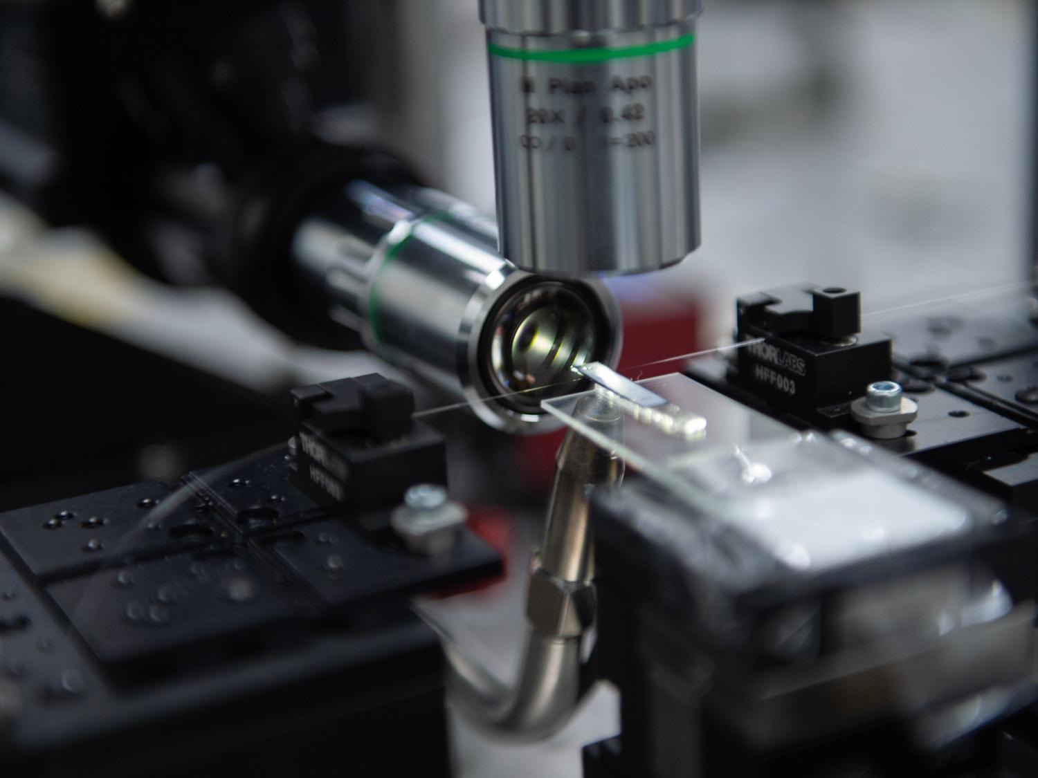 A glass rectangle sits at the forefront, slightly out of focus, touching a hair-like structure stretched between two black clasps. Two silver cylinders point at the structure, one from above and one across from the glass rectangle. 