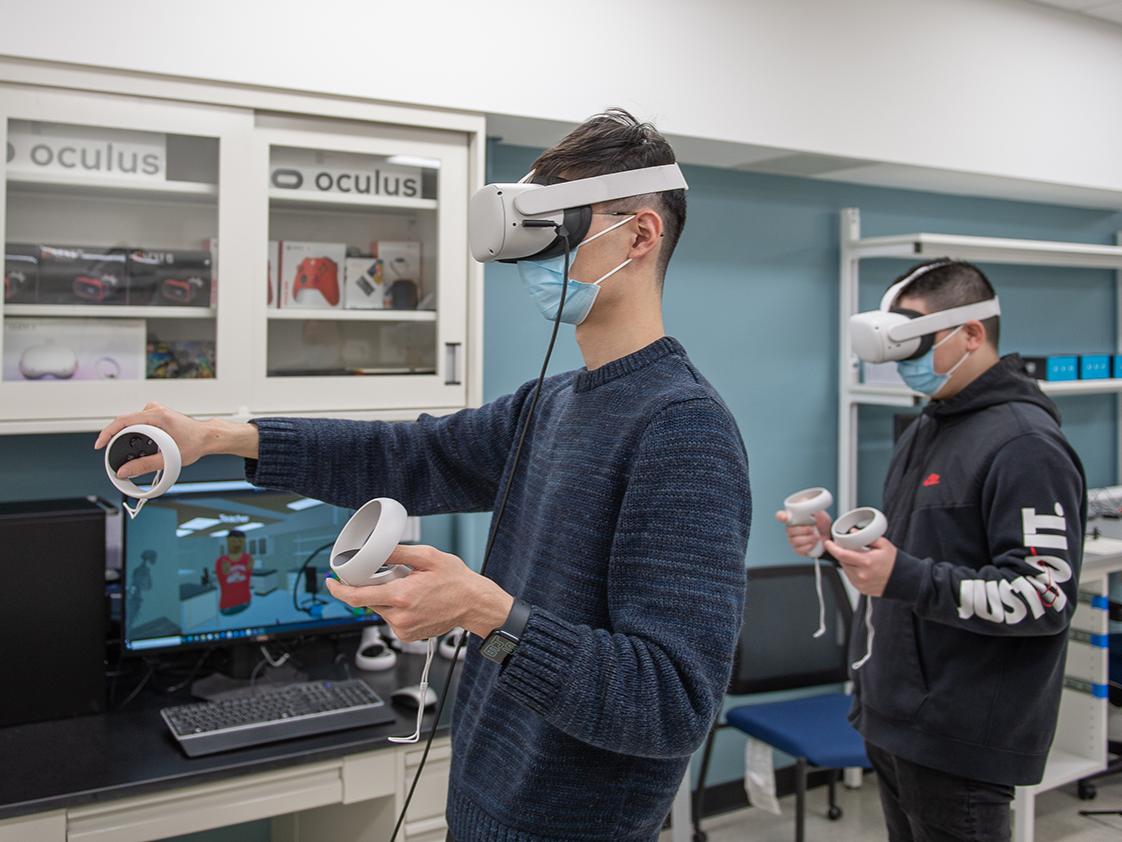 Two men stand while using virtual reality headsets and hand controls. 