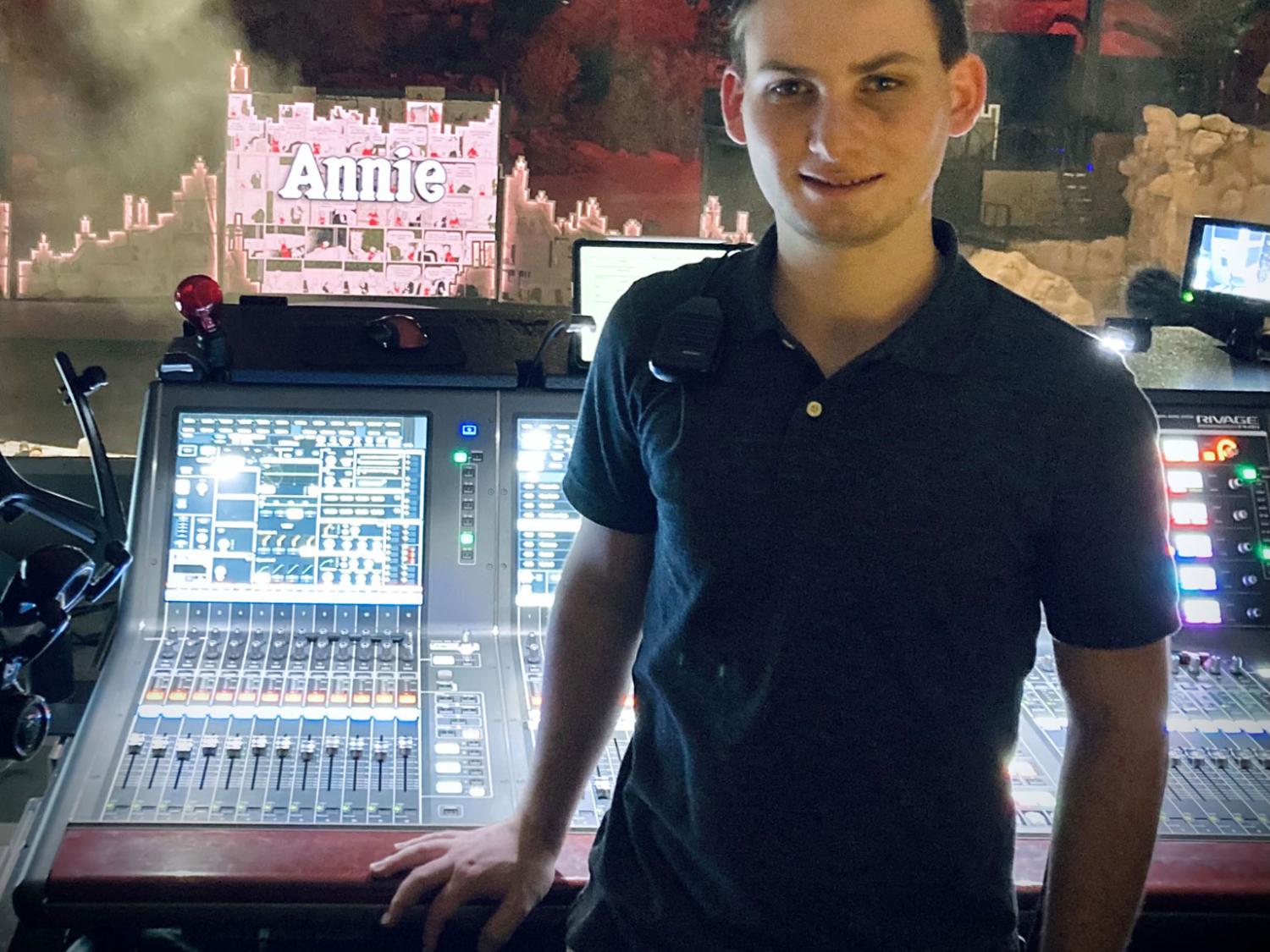 A young man stands in front of a sound production board with its knobs and buttons.