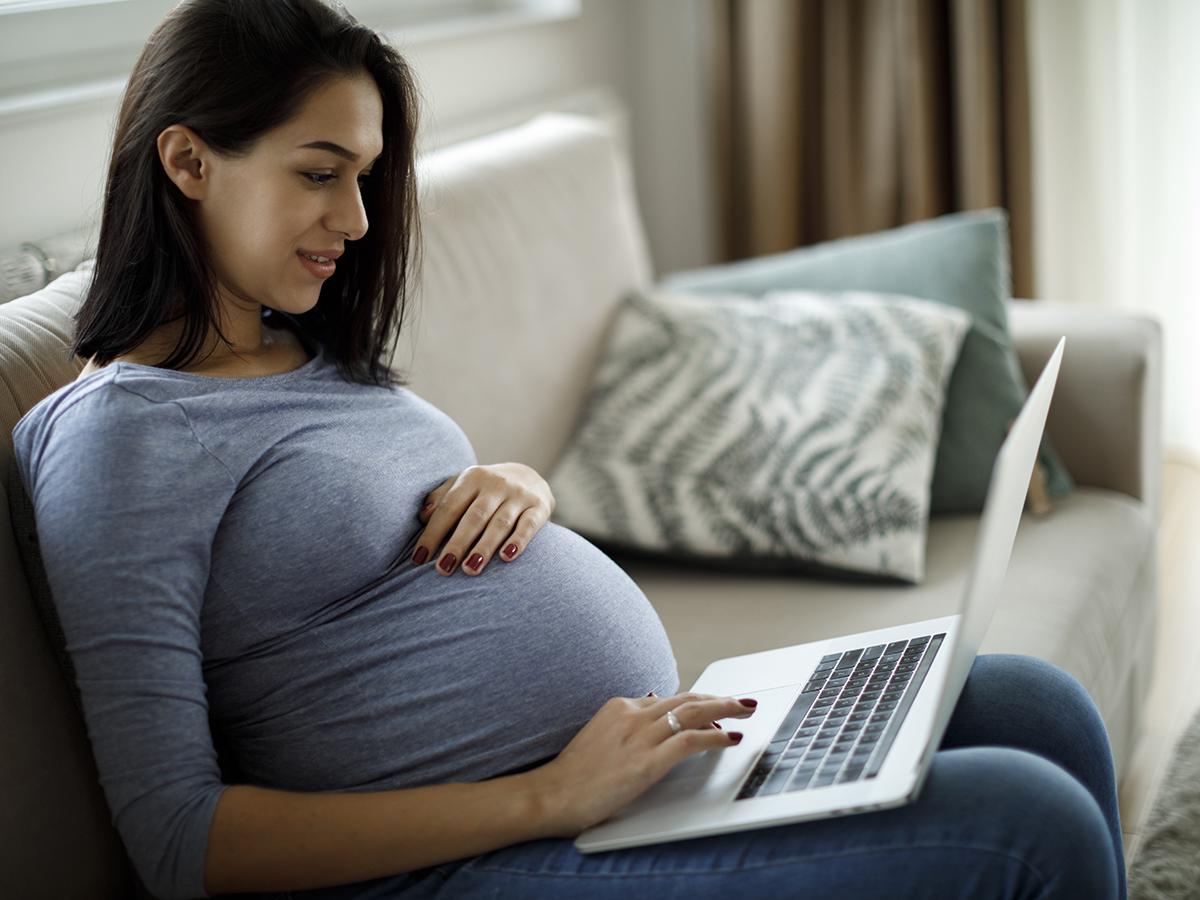 A pregnant woman holds her baby bump with one hand while typing on a laptop with her other hand while seated on a couch. 