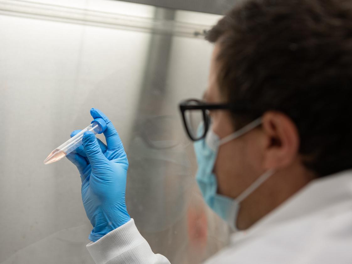 A researcher holds up a syringe containing a small amount of reddish translucent liquid.