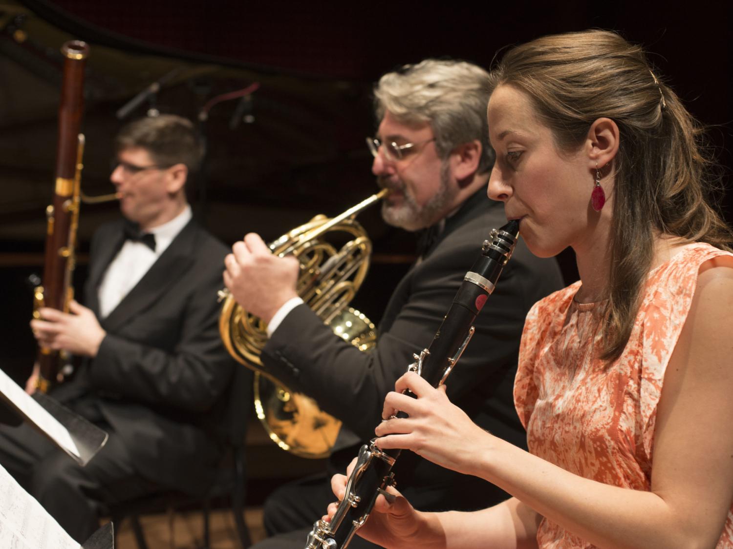 Three wind-instrument musicians sit at their stands and perform.