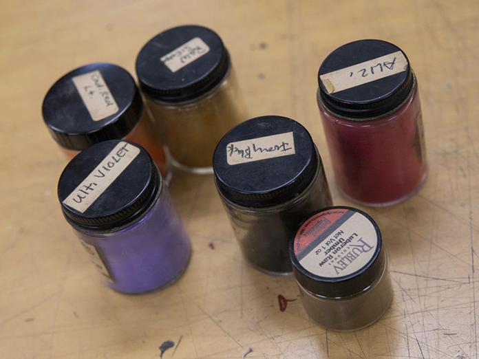 six small glass jars with black lids, each filled with a different color of paint.