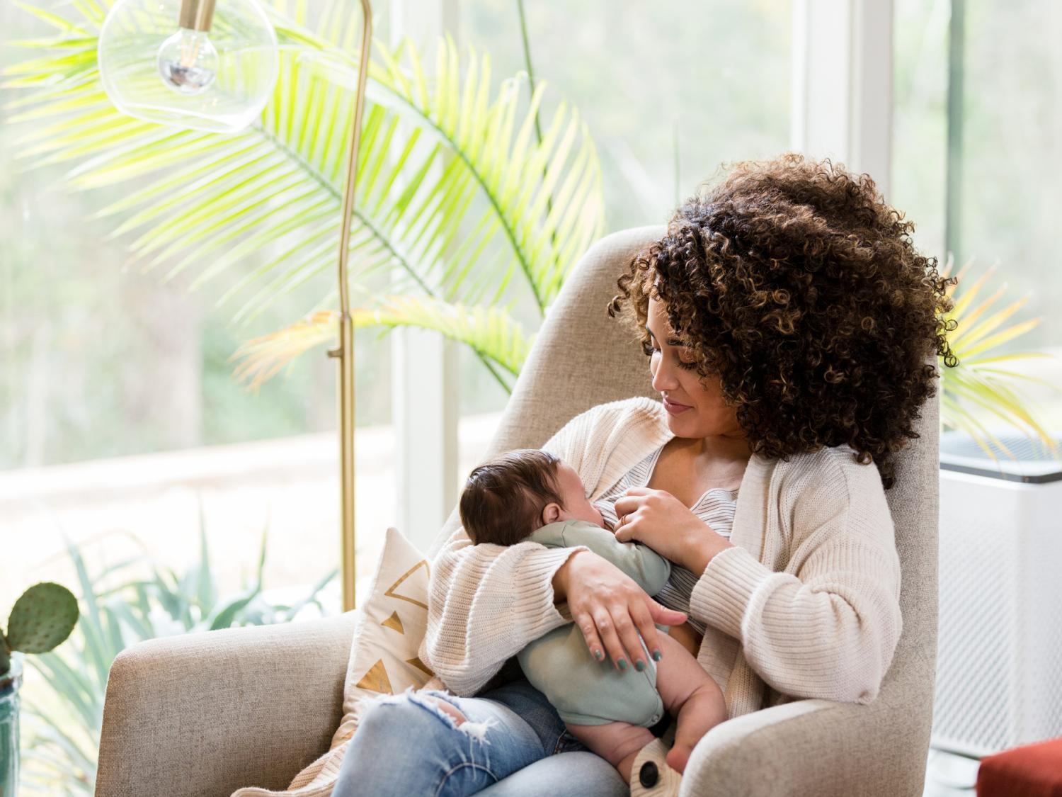 A woman sits in a chair, breastfeeding her baby. A lamp and some plants are in the background, as is a large window.
