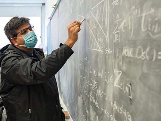 A man wearing a mask writes on a chalkboard.