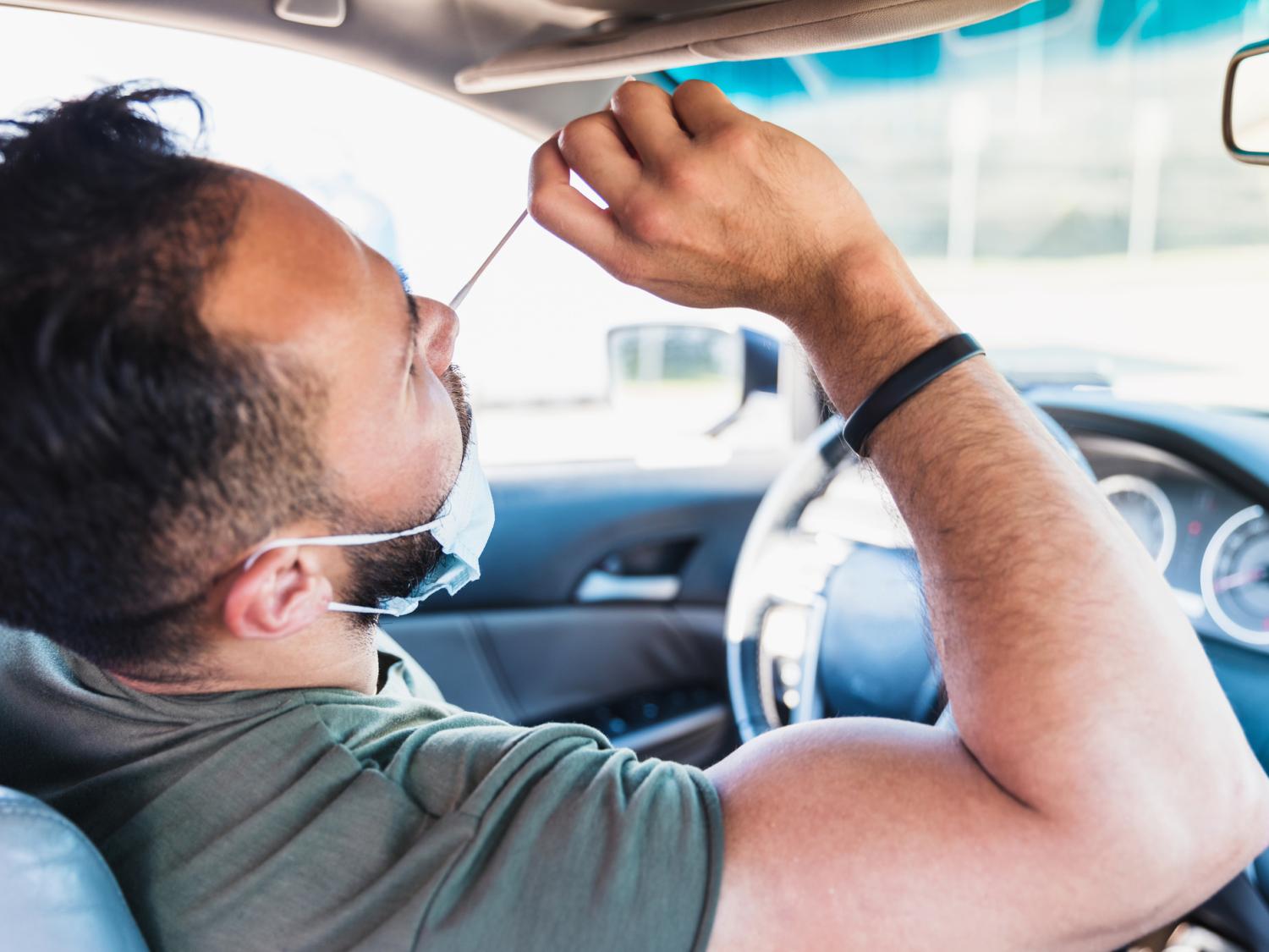 A man sitting in the driver’s seat of a car uses his right hand to insert a nasal swab into his nose, with his head tilted backward slightly. 