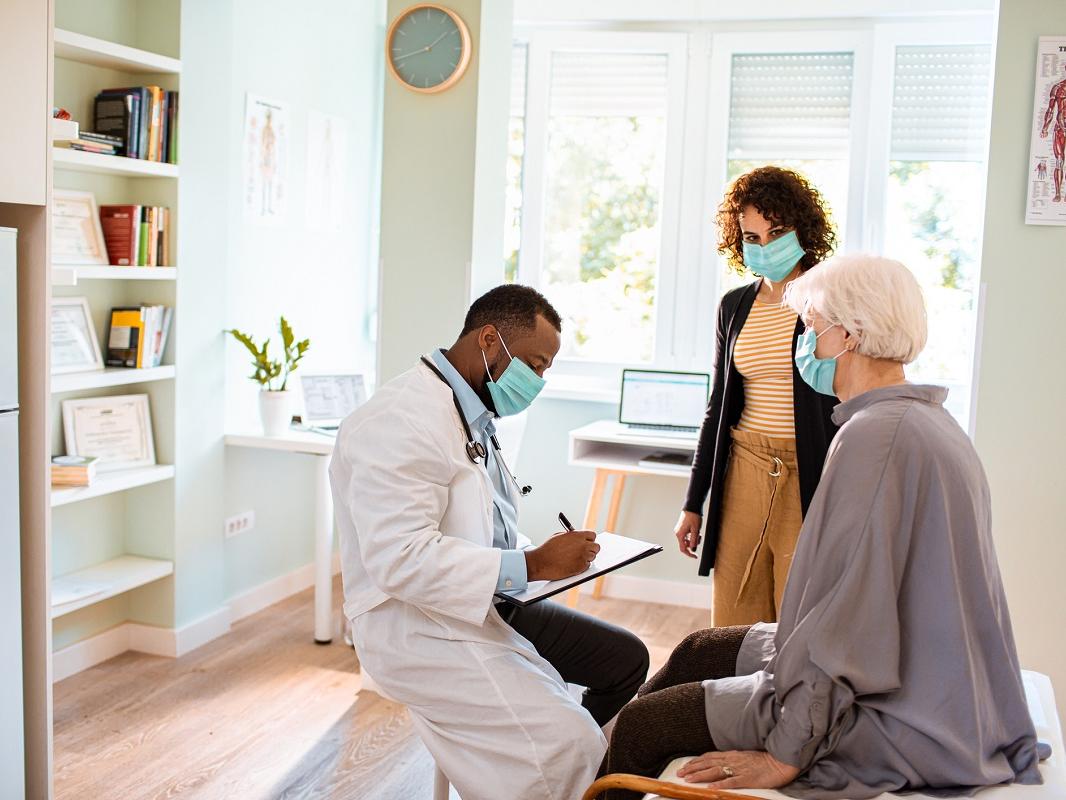 In a doctor’s office, a doctor is seated and writing in a chart while sitting across from a seated, elderly female patient. A younger woman stands by her side. All are wearing face masks.