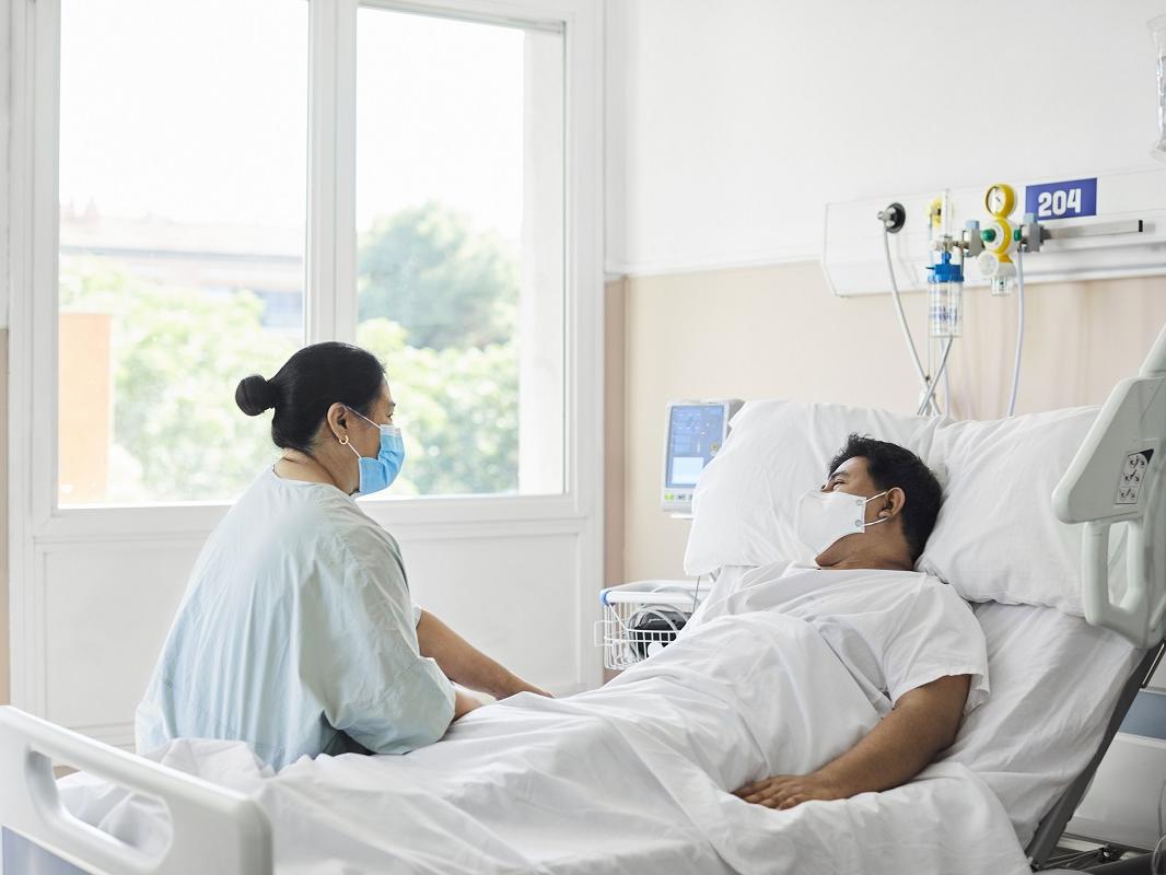 A woman sits at the edge of a hospital bed. A man lies in the bed. Both wear masks.