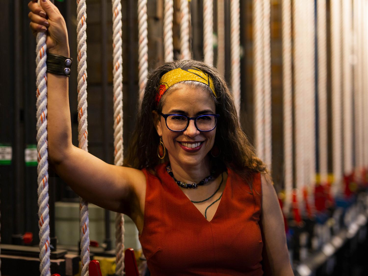 A woman wearing glasses and a hair sash stands on front of a line of pulley ropes.