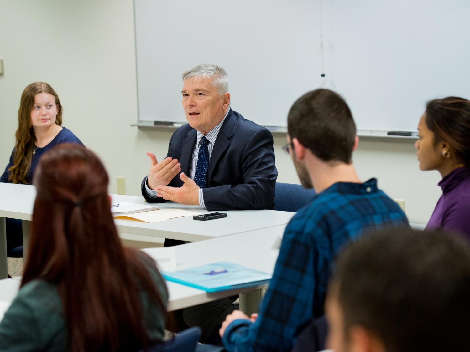Penn State President Eric Barron sits alongside students at the front of a classroom, where he is speaking and teaching class.
